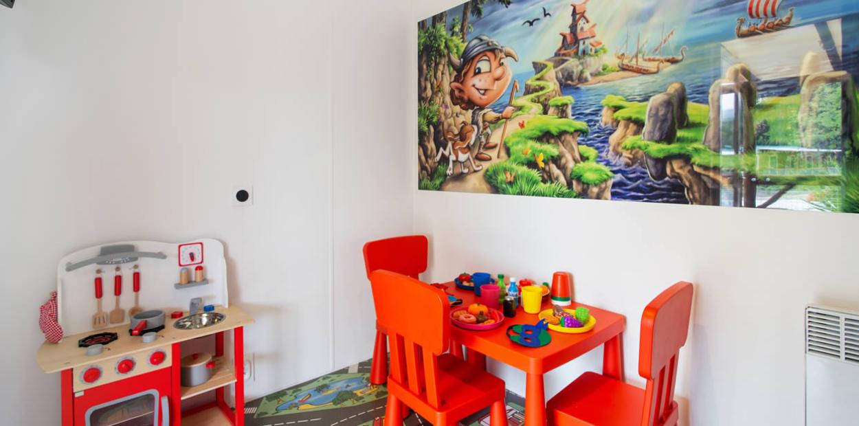 Angolo giochi per bambini con tavolo, sedie rosse e cucina giocattolo.