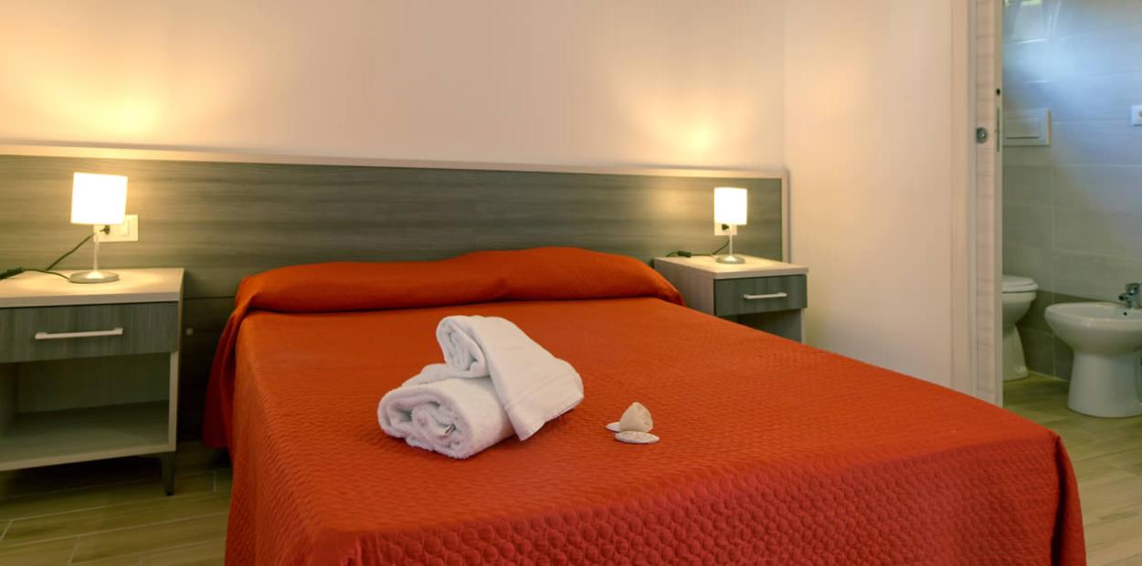 Camera d'albergo con letto matrimoniale, lenzuola rosse, bagno e luci soffuse.