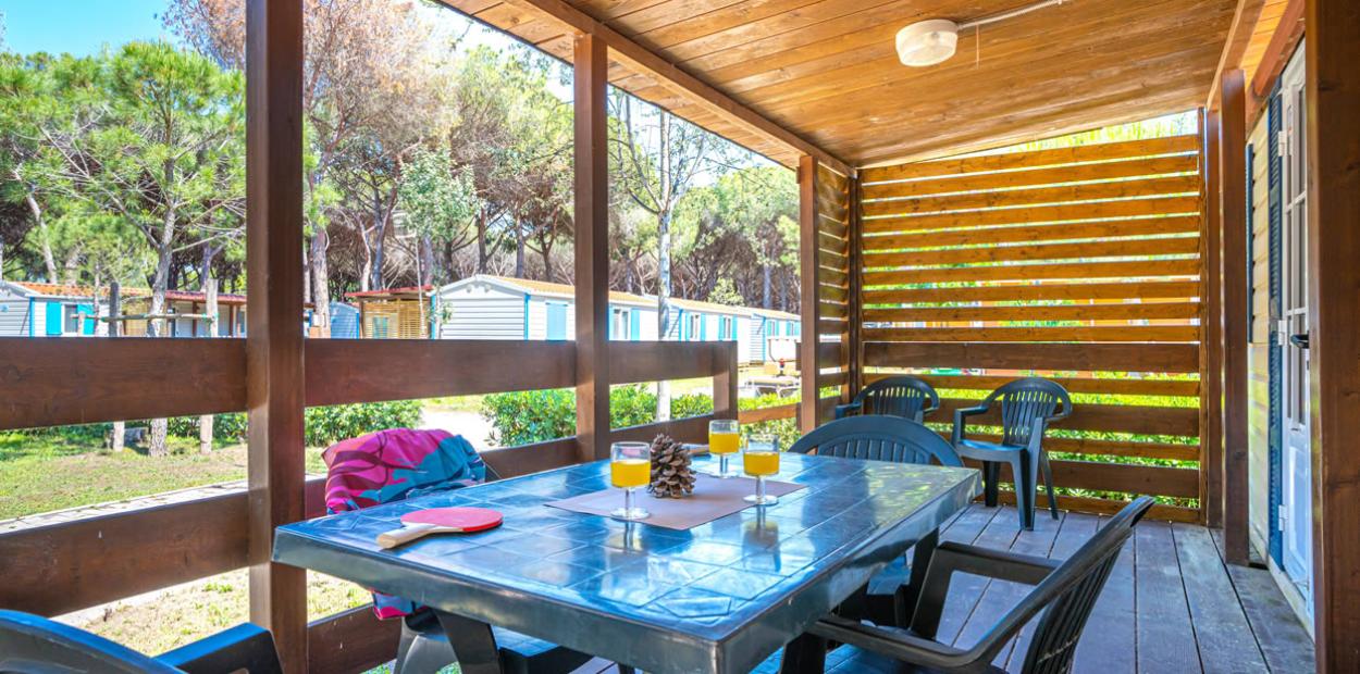 Veranda in legno con tavolo, sedie, bevande e vista su casette tra gli alberi.