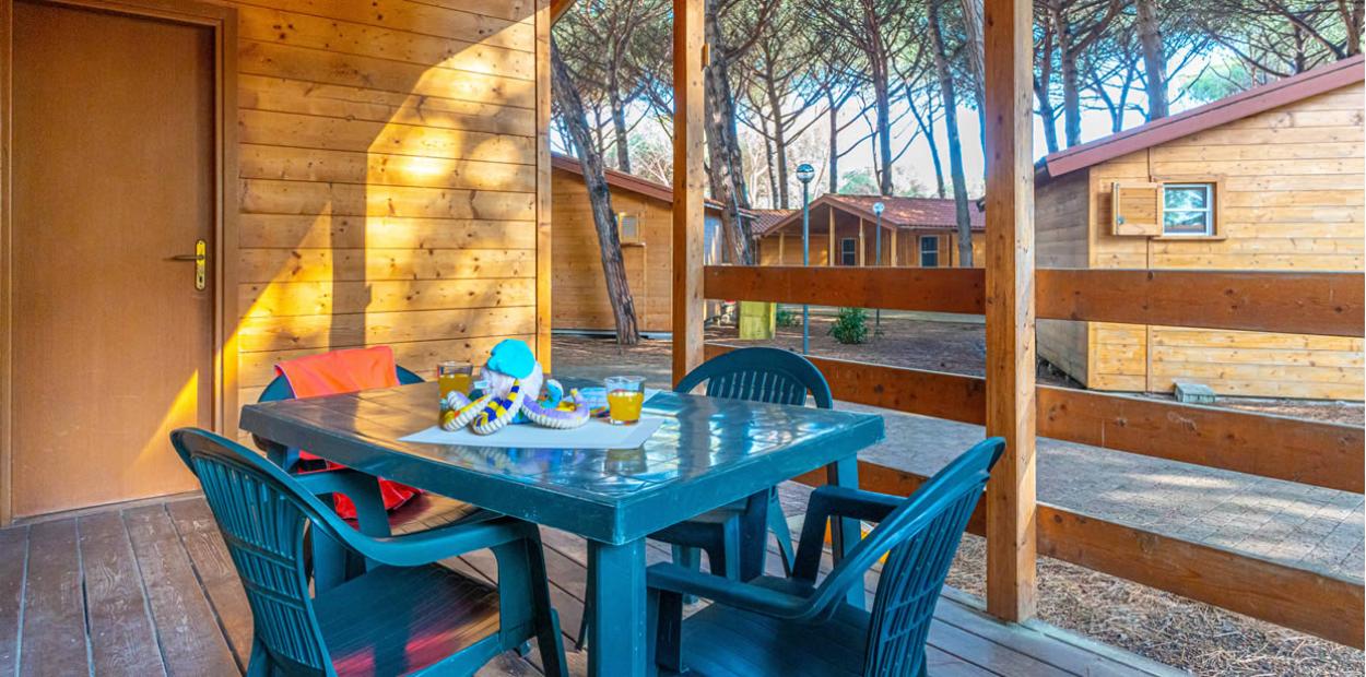 Veranda in legno con tavolo e sedie, immersa in un bosco di pini.