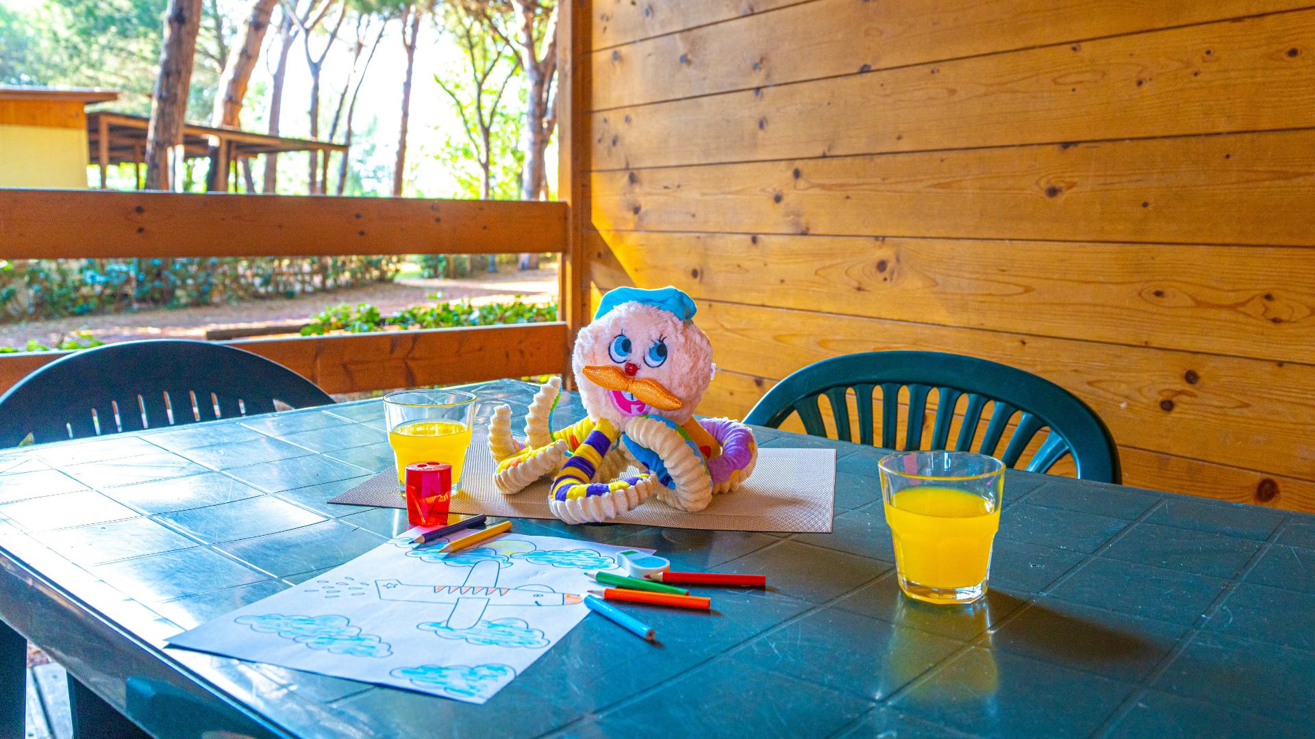 Tavolo con disegni, colori, peluche e bicchieri di succo d'arancia all'aperto.