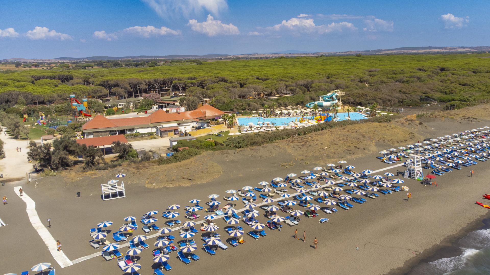 Spiaggia con ombrelloni, piscina e scivoli d'acqua, circondata da pineta.