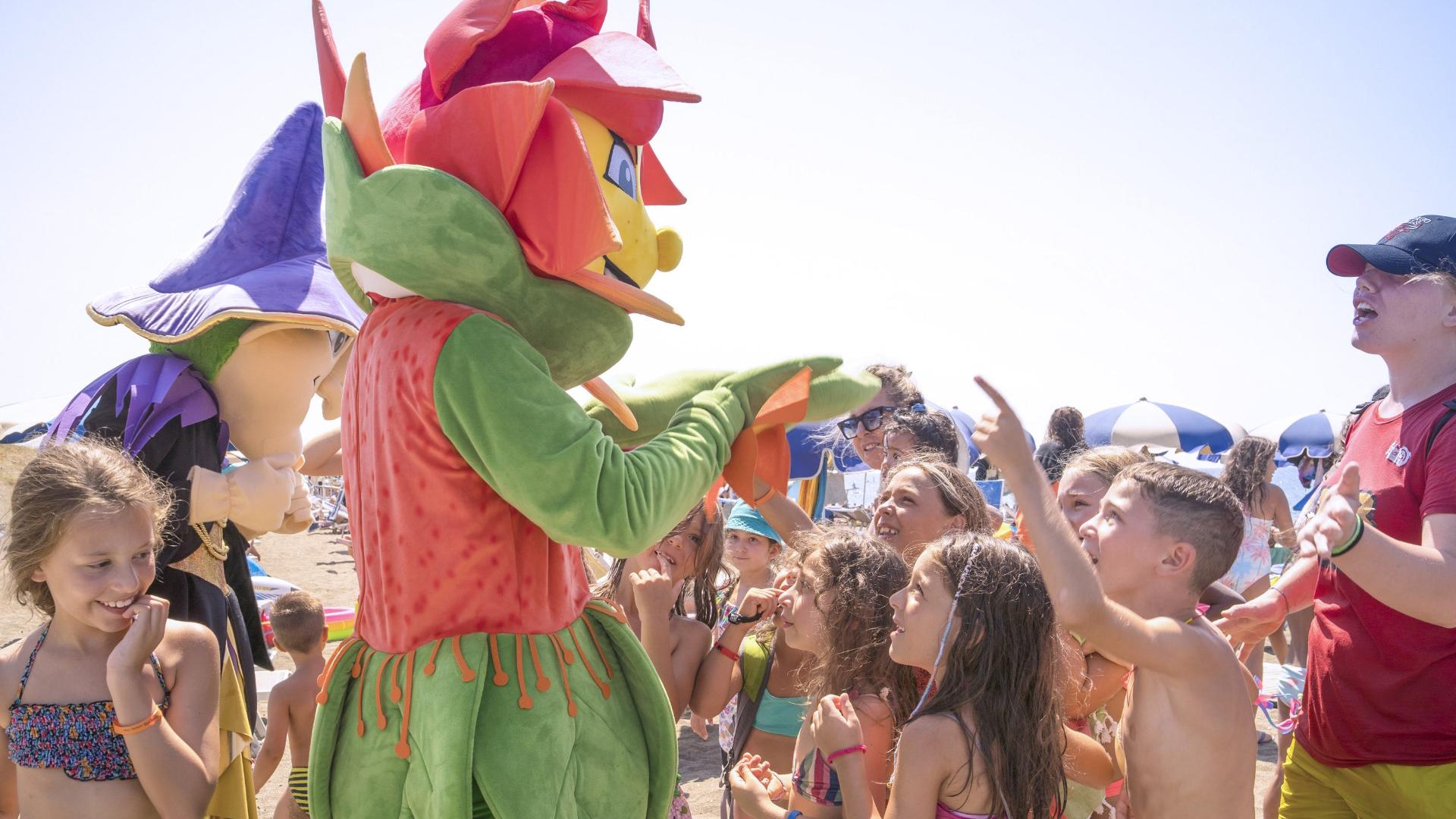 Personaggi in costume interagiscono con bambini sulla spiaggia in una giornata soleggiata.