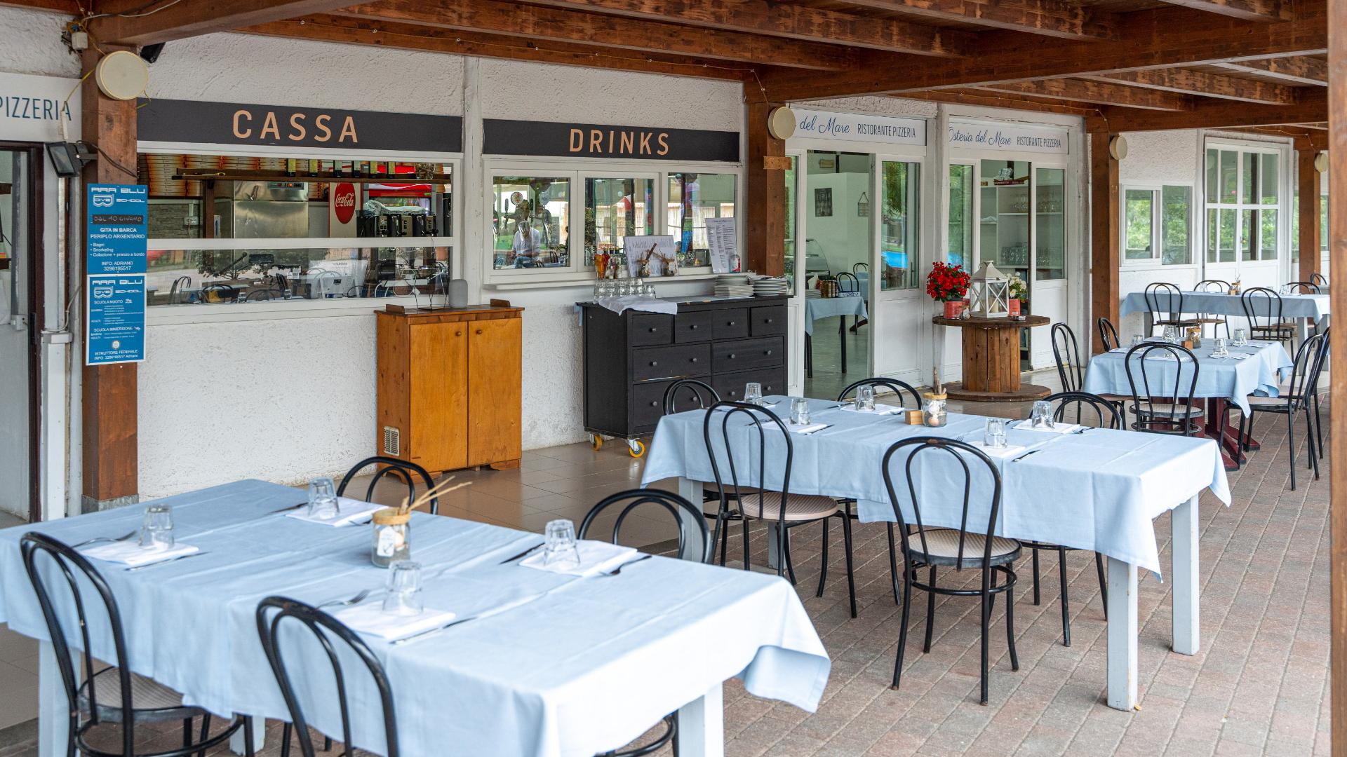Ristorante all'aperto con tavoli apparecchiati, insegna 'Cassa' e 'Drinks' visibile.