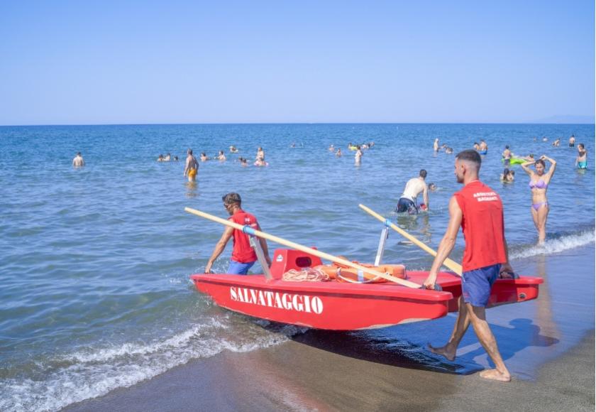 Bagnini portano un pattino di salvataggio rosso in mare, spiaggia affollata.