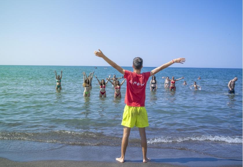 Istruttore guida esercizi di gruppo in mare, indossa maglietta rossa con scritto 'CREW'.
